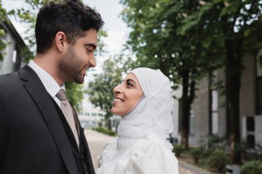 Beyaz tesettürlü mutlu Müslüman gelin ve takım elbiseli damat birbirine bakıyor.