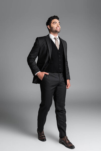 full length of stylish muslim groom in suit posing with hand in pocket while standing on grey