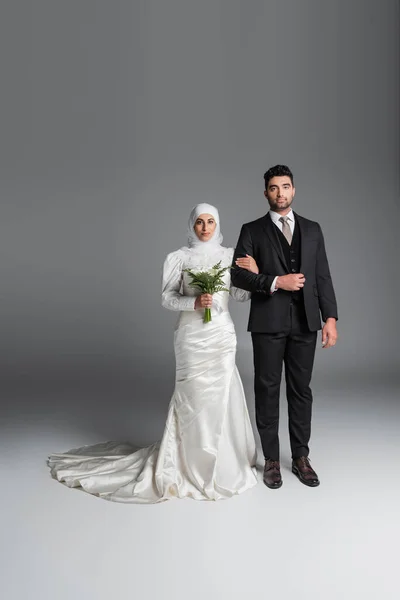 新郎の完全な長さスーツ姿でイスラム教徒の花嫁と一緒に立って結婚式の花束のカラユリの花を灰色 — ストック写真
