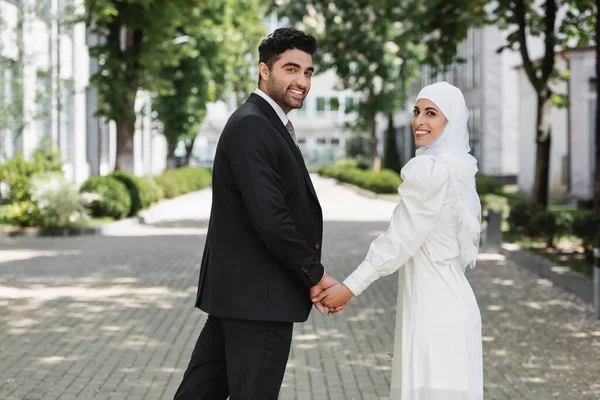 Mutlu damat, gülümseyen Müslüman gelinle el ele tutuşuyor ve birlikte yürüyor. 