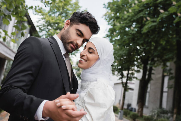 cheerful muslim newlyweds with closed eyes holding hands