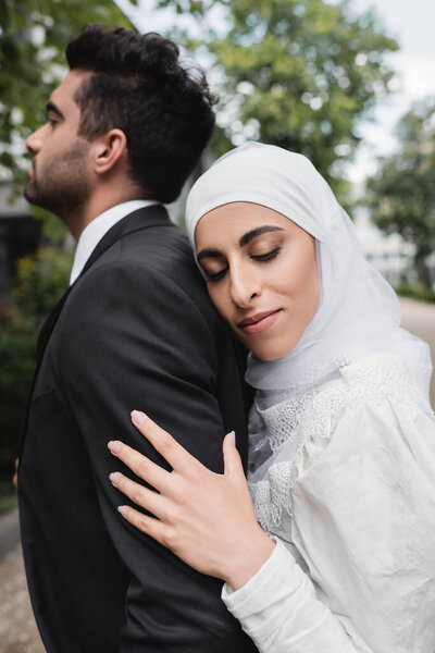 happy muslim bride in hijab and wedding dress hugging groom 