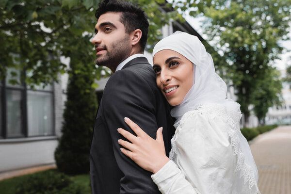 pleased muslim bride in hijab and wedding dress hugging groom 