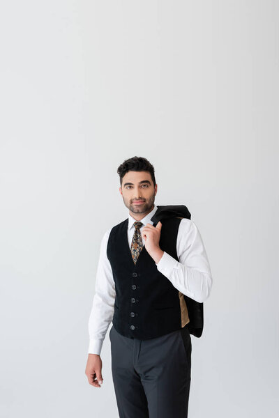 muslim man in vest holding blazer and looking at camera isolated on grey