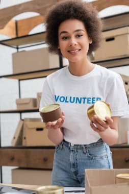 young african american volunteer with canned food smiling at camera in charity center