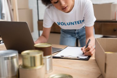 cropped view of african american volunteer writing on clipboard near laptop and blurred tins