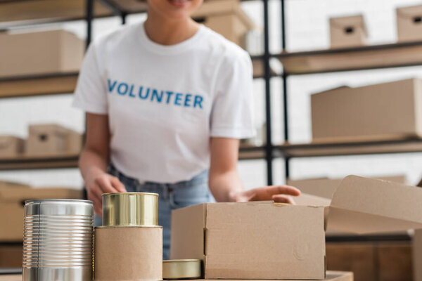 partial view of blurred african american woman working near canned foodstuff in volunteer center