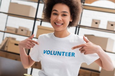 happy african american woman pointing at volunteer lettering on t-shirt in charity center