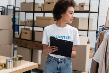 african american volunteer with clipboard looking at garments in charity center