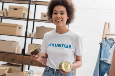 cheerful african american woman with canned food looking at camera in charity center