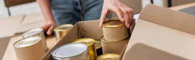 cropped view of african american volunteer packing canned food in box in charity storehouse, banner