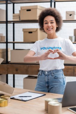 happy african american volunteer showing heart sign near clipboard on desk 