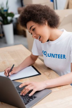 african american woman writing and talking on smartphone near laptop in volunteer center