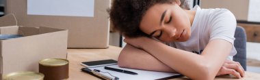 tired african american volunteer sleeping near smartphone and clipboard in charity warehouse, banner
