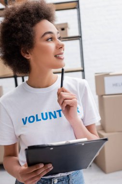 thoughtful african american volunteer with clipboard and pen looking away in charity warehouse
