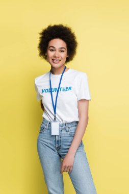 young african american volunteer with blank name tag standing with hand behind back isolated on yellow