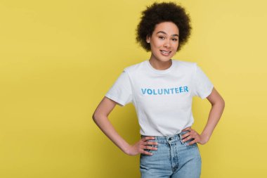 happy african american woman in t-shirt with volunteer lettering standing with hands on hips isolated on yellow