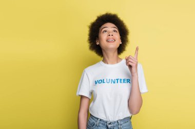 smiling african american volunteer pointing with finger while looking up isolated on yellow