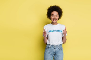 african american woman with volunteer inscription on t-shirt showing thumbs up isolated on yellow