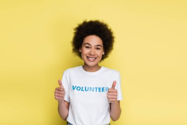cheerful african american volunteer showing thumbs up and looking at camera isolated on yellow