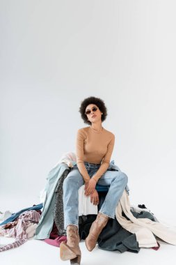 young african american woman sitting in sunglasses and jeans near mess of clothes on grey background
