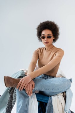 Kıvırcık Afrikalı, modaya uygun güneş gözlüklü Amerikalı kadın gri arka planda kıyafetlerin yanında oturuyor.