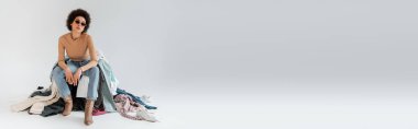 full length of fashionable african american woman sitting near plenty of garments on grey background, banner