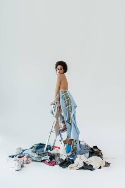 trendy african american woman standing on ladder near pile of clothes on grey background