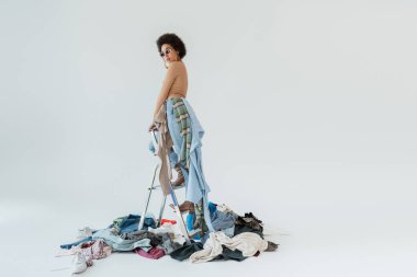fashionable african american woman standing on ladder above mess of garments on grey background