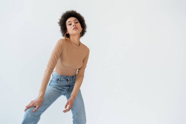 trendy african american woman in jeans and beige long sleeve shirt looking at camera isolated on grey