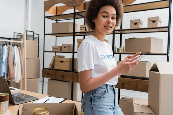 Positive African American Volunteer Using Smartphone Looking Camera Charity Warehouse — Zdjęcie stockowe