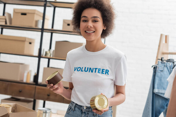 веселая африканская американка с консервами смотрит в камеру в благотворительном центре