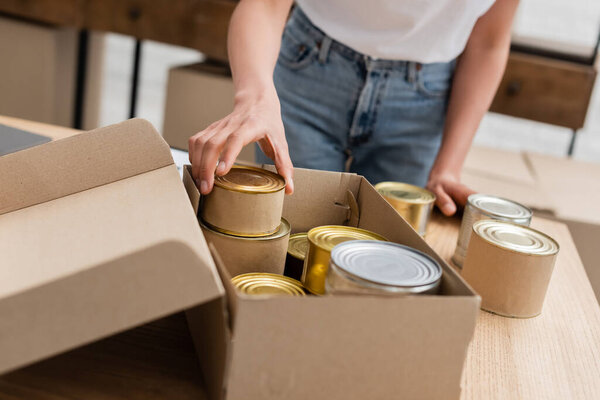 cropped view of african american woman packing canned food in box in volunteer center