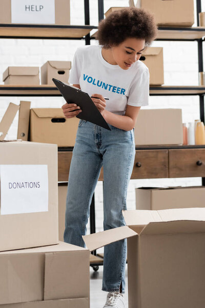 african american volunteer with clipboard working near carton boxes in charity warehouse