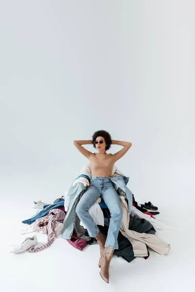 african american woman with hands behind head sitting near mess of clothes on grey background