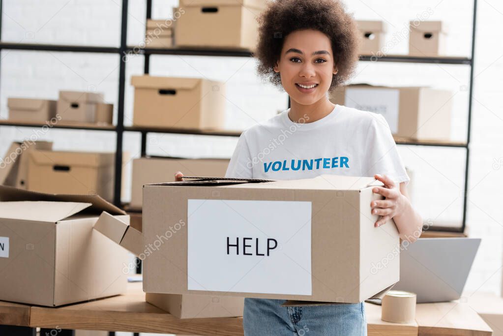 happy african american volunteer holding carton box with help lettering in charity center