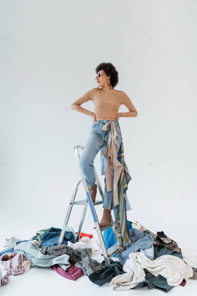 african american woman with hands on waist standing on ladder near mess of garments on grey background