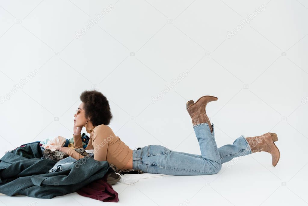 side view of african american woman in jeans and cowboy boots lying near pile of clothing on grey background