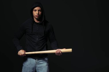 African american man in hoodie holding baseball bat and looking at camera isolated on black  clipart