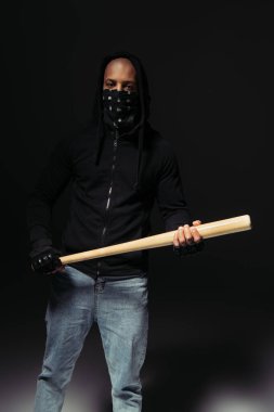 African american hooligan with covered face holding baseball bat on black background  clipart