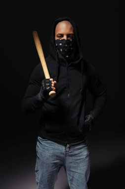 African american hooligan with scarf on face holding baseball bat on black background 