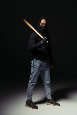 Full length of afrcan american hooligan in mask on face holding baseball bat on black background 