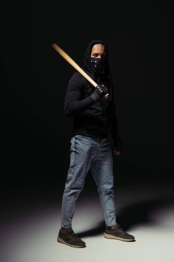 Full length of afrcan american hooligan in mask on face holding baseball bat on black background  clipart