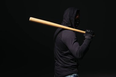 Side view of african american hooligan in mask and glove holding baseball bat isolated on black  clipart
