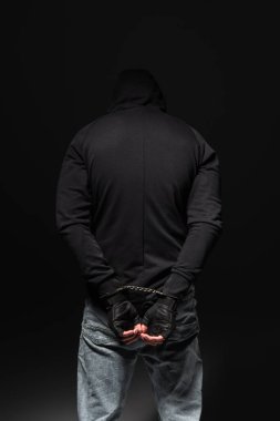 Back view of african american bandit in hoodie and handcuffs standing isolated on black  clipart