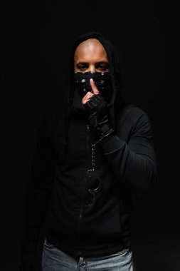 African american bandist in mask on face and handcuffs showing secret gesture isolated on black 