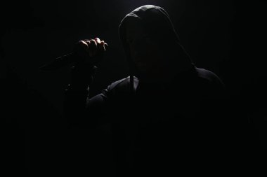 Silhouette of bandit in hoodie holding knife isolated on black  clipart