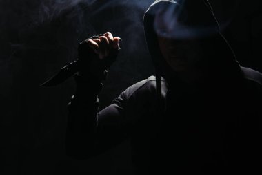Silhouette of african american bandit in hood holding knife on black background with smoke 
