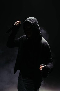 Silhouette of african american man in hood and gloves holding knife on black background with smoke  clipart