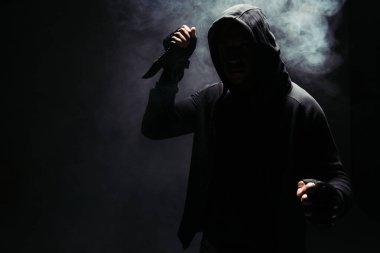 Silhouette of african american bandit in hoodie holding knife on black background with smoke  clipart